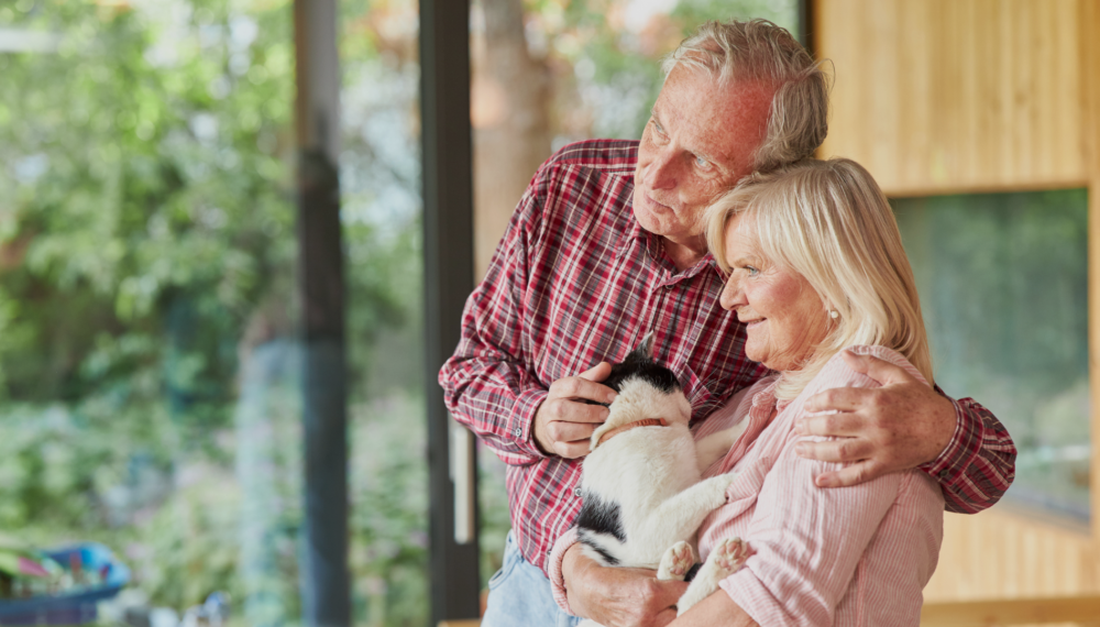 mature couple hugging, holding cat, and looking out the window smiling